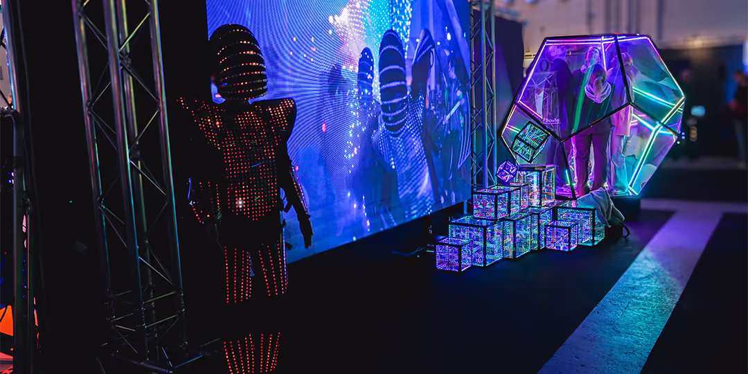 LLB Expo Varför ställa ut LED-klädd performer vid stor skärm; till höger en neonbelyst installation med genomskinlig polyeder och staplade ljuskuber, med besökare inuti, på mässgolvet.