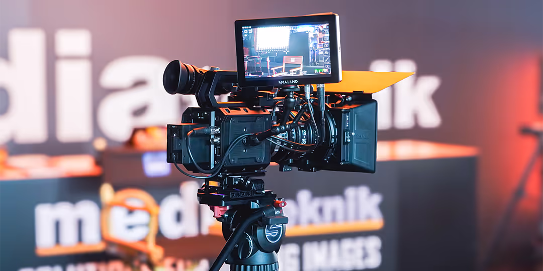 LLB Expo Varför ställa ut Professionell videokamera på stativ med extern skärm som visar livebild, mot oskarp mässmonter i bakgrunden.